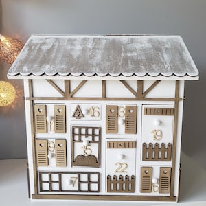 May include: A white wooden advent calendar in the shape of a house with a grey roof. It features numerous small drawers and doors, each numbered. The trim is brown, and the design has a rustic aesthetic.