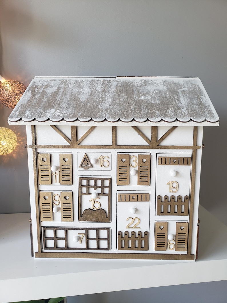 May include: A white wooden advent calendar in the shape of a house with a grey roof. It features numerous small drawers and doors, each numbered. The trim is brown, and the design has a rustic aesthetic.