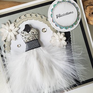 May include: A white card with a black and silver border. The card features a white feather boa dress with a black and white geometric pattern and silver glitter. The card also has a white flower and a circle with the word "F&eacute;licitations" in black text.