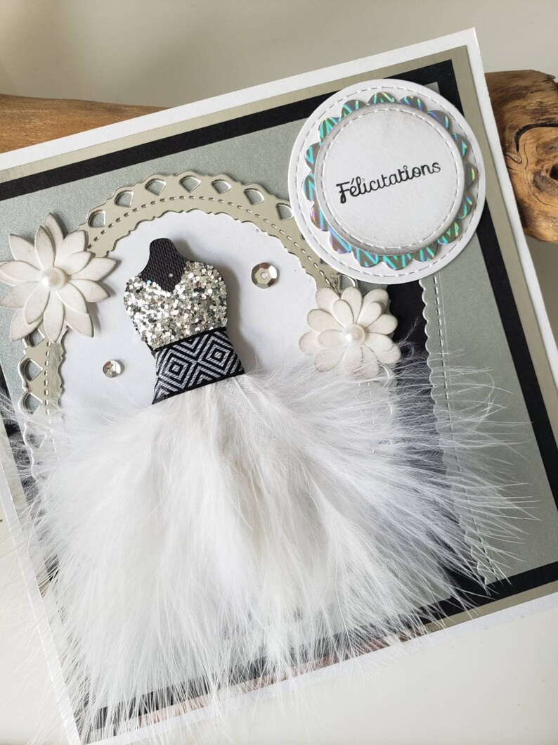 May include: A white card with a black and silver border. The card features a white feather boa dress with a black and white geometric pattern and silver glitter. The card also has a white flower and a circle with the word "F&eacute;licitations" in black text.