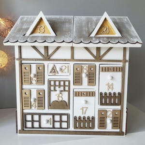 May include: A white wooden advent calendar shaped like a house, featuring a grey roof and gold detailing. The house has numerous drawers, each marked with numbers and decorative features such as a fence and shutters. Numbers 13 and 24 are displayed on the roof.