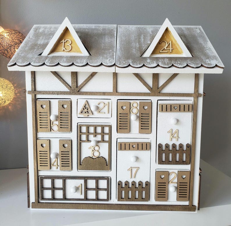 May include: A white wooden advent calendar shaped like a house, featuring a grey roof and gold detailing. The house has numerous drawers, each marked with numbers and decorative features such as a fence and shutters. Numbers 13 and 24 are displayed on the roof.