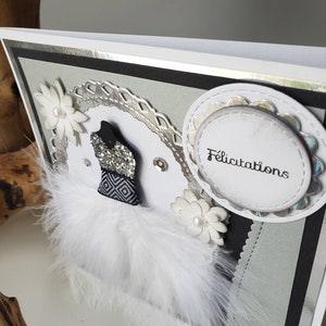 May include: A white card with a silver border and a black and white design. The card has a white feather trim, a silver and white flower, and a white circle with the word "Felicitation" in black text.