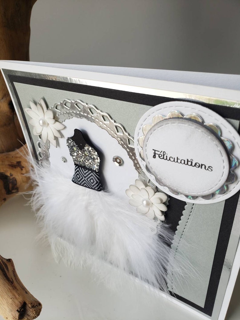 May include: A white card with a silver border and a black and white design. The card has a white feather trim, a silver and white flower, and a white circle with the word "Felicitation" in black text.