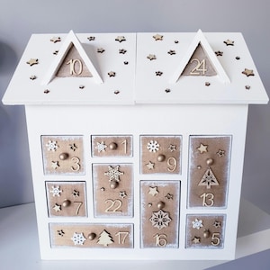 May include: A white wooden advent calendar shaped like a house. The roof features the numbers 10 and 24. The drawers are numbered 1 to 24 and decorated with wooden stars, snowflakes, and Christmas trees.