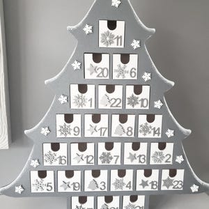 May include: A grey wooden Christmas tree advent calendar with 24 numbered drawers. Each drawer is decorated with a snowflake or star. The tree has white snowflake accents.