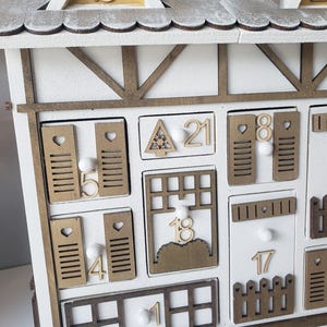 May include: A white wooden advent calendar shaped like a house. The roof is white with gold accents and numbers. The calendar features small doors and windows, each with a number from 1 to 24. The measurements are in centimeters.