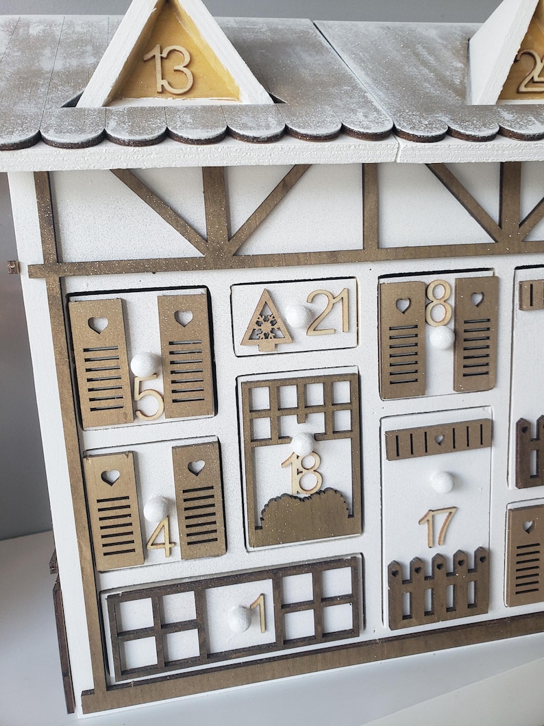 May include: A white wooden advent calendar shaped like a house. The roof is white with gold accents and numbers. The calendar features small doors and windows, each with a number from 1 to 24. The measurements are in centimeters.