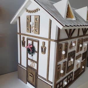 May include: A wooden advent calendar shaped like a house, painted white with brown trim. It features numerous small doors and windows, each marked with a number. The roof is white with a silver glitter effect. A small wreath and the number 20 are on the front.