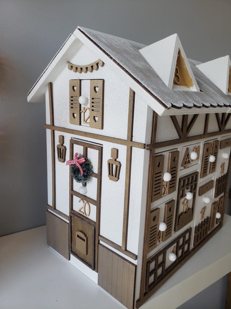 May include: A wooden advent calendar shaped like a house, painted white with brown trim. It features numerous small doors and windows, each marked with a number. The roof is white with a silver glitter effect. A small wreath and the number 20 are on the front.
