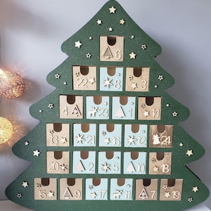 May include: A green wooden Christmas tree advent calendar with 24 small drawers. Each drawer is numbered and decorated with wooden cutouts of stars, snowflakes, and festive shapes. The tree is decorated with small star cutouts.