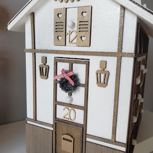 May include: A festive white and brown house with gold detailing, perfect for the holidays. It features a small wreath, the number 20, and a mailbox. The roof has a garland of lights and the number 12 is visible.