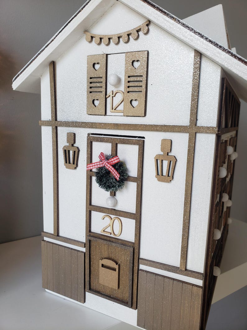May include: A festive white and brown house with gold detailing, perfect for the holidays. It features a small wreath, the number 20, and a mailbox. The roof has a garland of lights and the number 12 is visible.