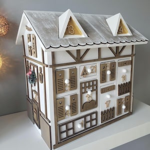 May include: A white wooden advent calendar in the shape of a house, featuring a gray roof and dormers. It has numerous small doors and windows, each marked with a number. A small Christmas wreath is attached.