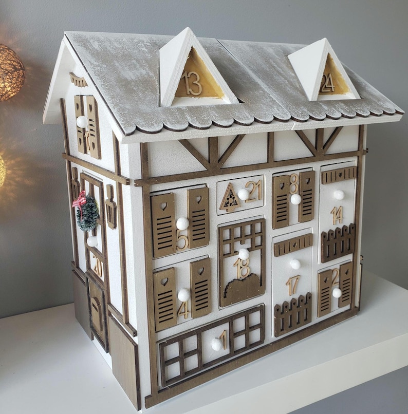 May include: A white wooden advent calendar in the shape of a house, featuring a gray roof and dormers. It has numerous small doors and windows, each marked with a number. A small Christmas wreath is attached.