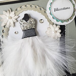 May include: A white feather boa with a silver and black sequined bodice and a black and white geometric pattern. The boa is on a silver and black card with the word "Felicitation" in a circle.