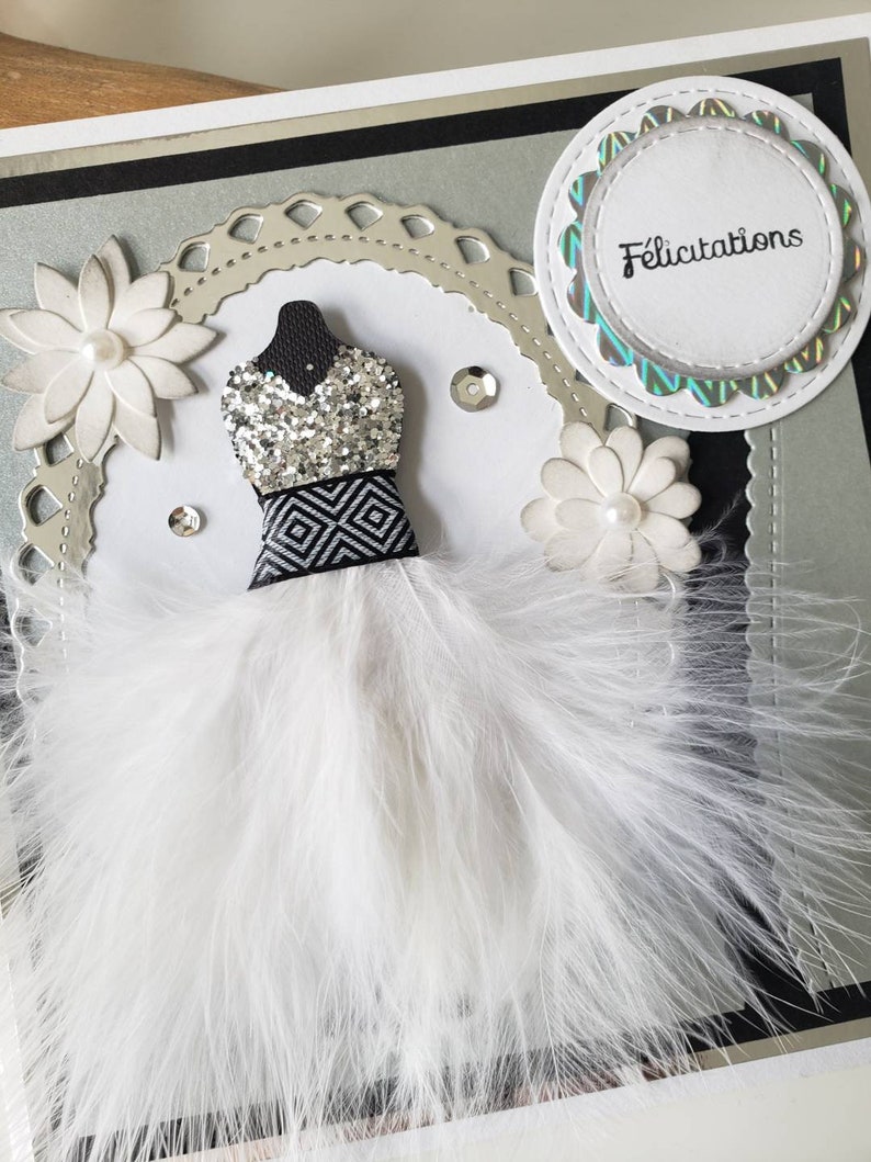 May include: A white feather boa with a silver and black sequined bodice and a black and white geometric pattern. The boa is on a silver and black card with the word "Felicitation" in a circle.