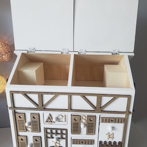 May include: A white wooden advent calendar with a decorative brown trim and multiple small drawers. The calendar has two hinged top doors and is decorated with numbers and small wooden cutouts.