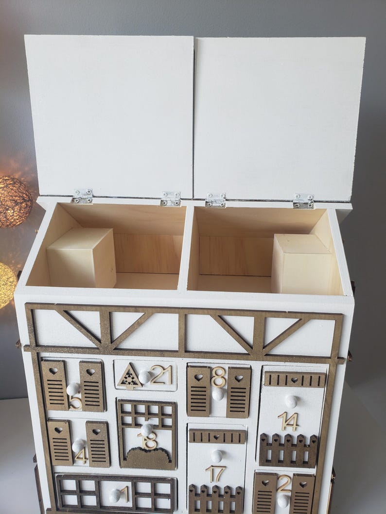 May include: A white wooden advent calendar with a decorative brown trim and multiple small drawers. The calendar has two hinged top doors and is decorated with numbers and small wooden cutouts.