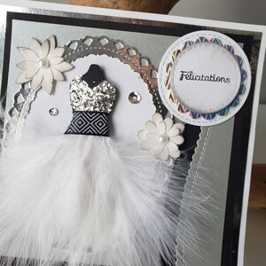May include: A silver and black card with a white feather boa, a silver glitter dress, and a white flower. The card has a circle with the word "Felicitation" in black text.