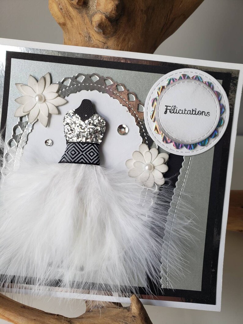 May include: A silver and black card with a white feather boa, a silver glitter dress, and a white flower. The card has a circle with the word "Felicitation" in black text.