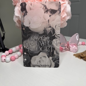 Puede incluir: Un inserto de planificador rectangular transparente con un dise&ntilde;o floral en blanco y negro. El texto "Stay Cute & Stay On Budget" se muestra en el inserto. Flores rosas y cuentas decorativas son visibles.