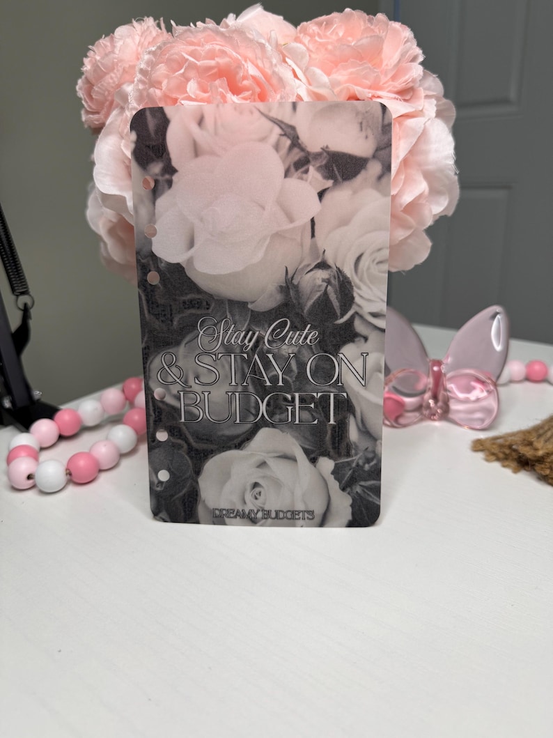 Puede incluir: Un inserto de planificador rectangular transparente con un dise&ntilde;o floral en blanco y negro. El texto "Stay Cute & Stay On Budget" se muestra en el inserto. Flores rosas y cuentas decorativas son visibles.