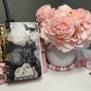 Puede incluir: Una agenda rosa con detalles dorados y una cubierta con un dise&ntilde;o floral que dice "Stay Cute & Stay on Budget". Un ramo de flores rosas en un jarr&oacute;n blanco est&aacute; junto a la agenda. Cuentas decorativas y una mariposa tambi&eacute;n est&aacute;n presentes.