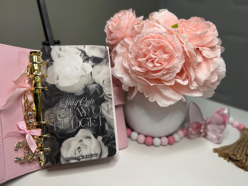 Puede incluir: Una agenda rosa con detalles dorados y una cubierta con un dise&ntilde;o floral que dice "Stay Cute & Stay on Budget". Un ramo de flores rosas en un jarr&oacute;n blanco est&aacute; junto a la agenda. Cuentas decorativas y una mariposa tambi&eacute;n est&aacute;n presentes.