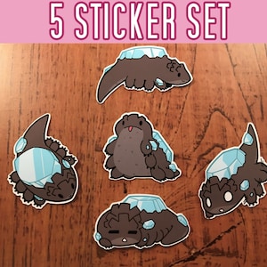 May include: Set of five stickers featuring cartoon illustrations of brown and black creatures with blue and white crystal-like shells. The stickers are on a wooden surface.  "5 Sticker Set" is written in pink at the top of the image.