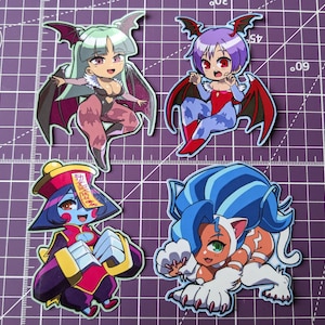 May include: Four colorful anime character stickers. The stickers feature a purple-haired vampire girl, a blue-haired cat girl, a green-haired vampire girl, and a red-haired girl in a red and gold outfit. The stickers are all in a chibi style.
