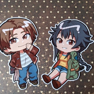May include: Two anime-style chibi character stickers. The character on the left is a boy with brown hair and a brown jacket. The character on the right is a girl with black hair and a green jacket. Both characters are smiling.