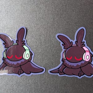 Op de afbeelding: Twee stickers met een cartoonmot met grote oren, met een koptelefoon op. Eén mot heeft mintgroene koptelefoon, de andere roze. Beide hebben rode ogen en een paarse omtrek.