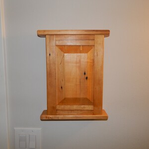 May include: A small wooden cabinet with a single door and a shelf. The cabinet is made of light wood with a natural finish.