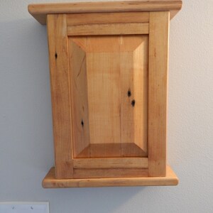 May include: A small wooden cabinet with a single door and a simple design. The cabinet is made of light wood with a natural finish. It is mounted on a wall.