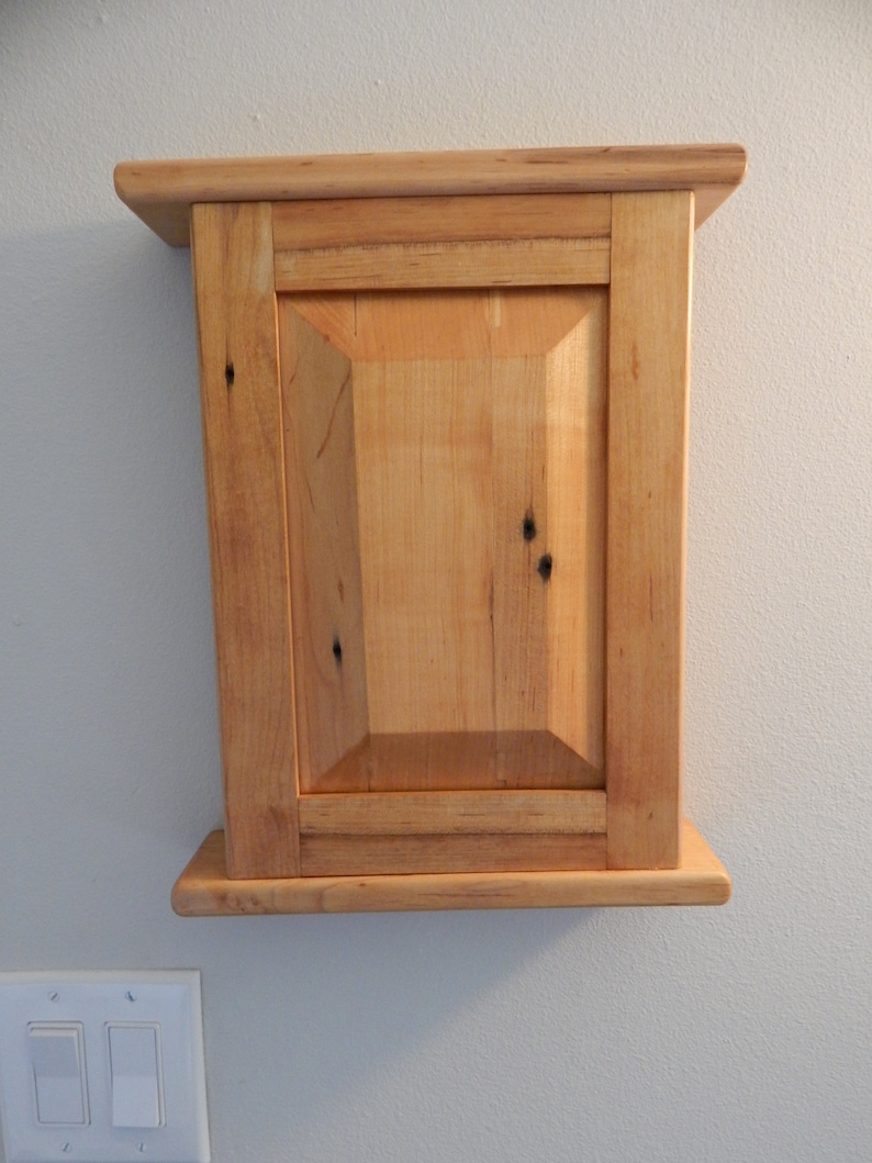 May include: A small wooden cabinet with a single door and a simple design. The cabinet is made of light wood with a natural finish. It is mounted on a wall.