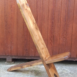 May include: A wooden folding chair with a simple design. The chair is made of natural wood and has a rustic look. The chair is in a folded position, with the back legs crossed over the front legs.