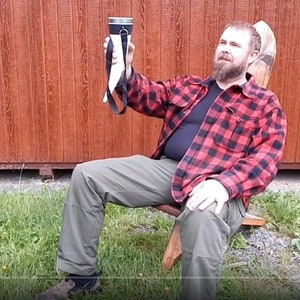 May include: A man wearing a red and black plaid shirt sits on a wooden chair and holds up a white drinking horn with a black strap. The horn has a metal top and is filled with a liquid.