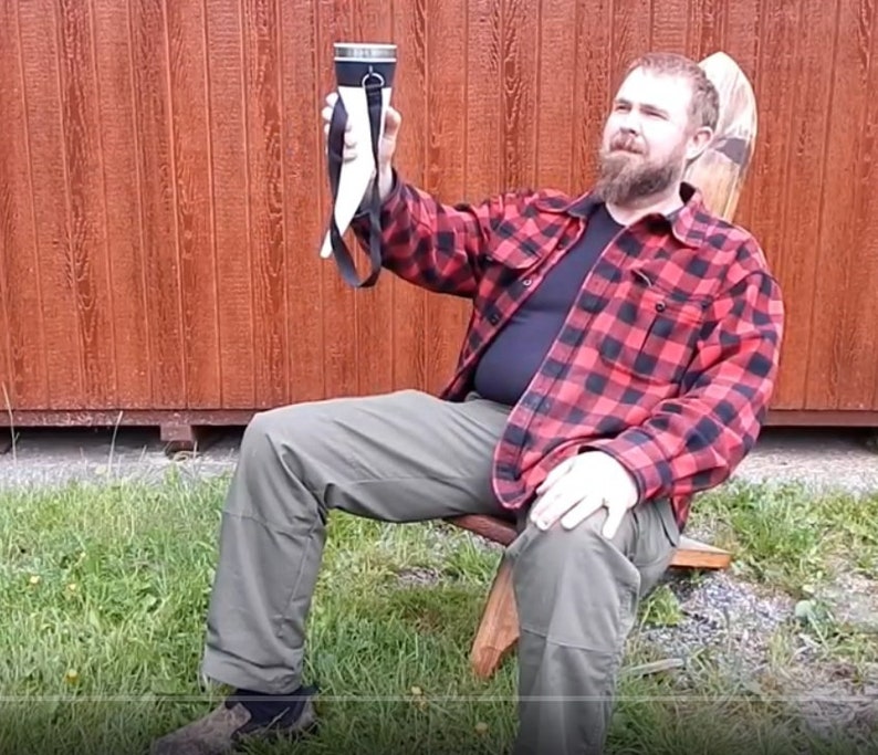 May include: A man wearing a red and black plaid shirt sits on a wooden chair and holds up a white drinking horn with a black strap. The horn has a metal top and is filled with a liquid.