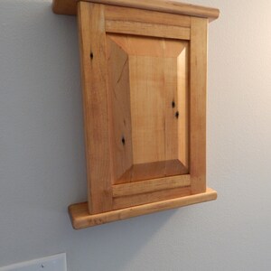 May include: A wooden cabinet with a single door and a shelf. The cabinet is made of light wood with a natural finish. It is mounted on a wall.