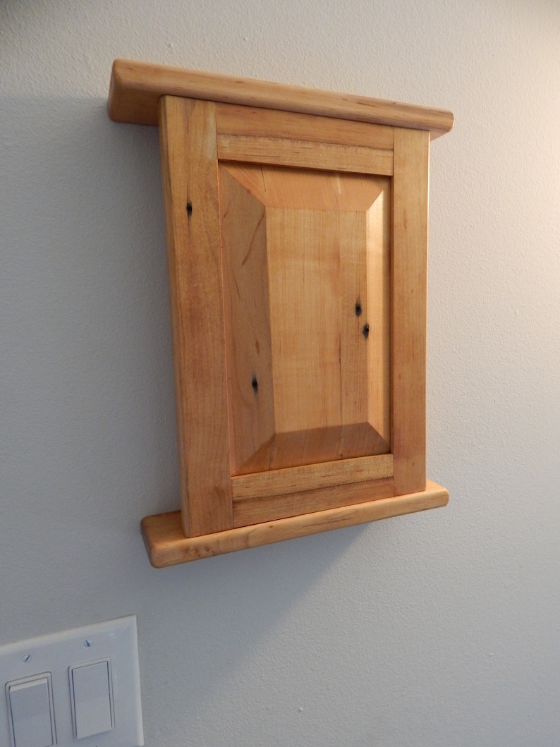 May include: A wooden cabinet with a single door and a shelf. The cabinet is made of light wood with a natural finish. It is mounted on a wall.