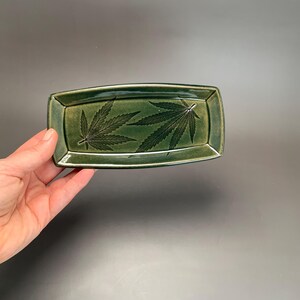 May include: A rectangular, green ceramic tray with a glossy finish. The tray features two detailed cannabis leaf designs in a darker green. The edges are slightly raised, and the tray is held by a hand against a dark background.