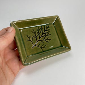 May include: A small, rectangular, olive-green ceramic dish with a black, stylized fern design. The dish has slightly raised edges and a glossy finish. The handmade pottery piece is held in a hand against a white background.