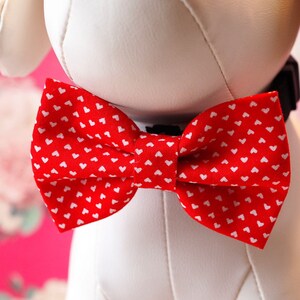 May include: A white stuffed dog wearing a red bow tie with white hearts. The bow tie is tied around the dog's neck.
