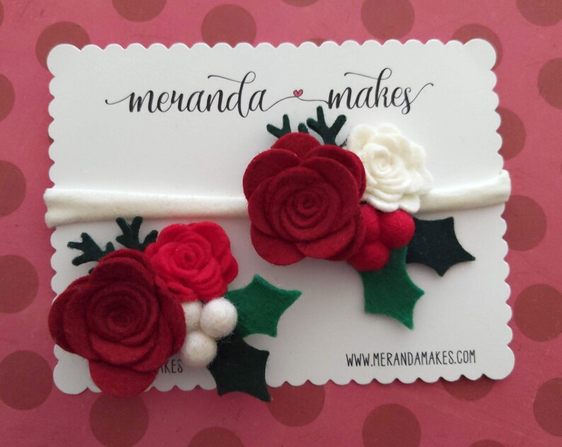 May include: Two felt Christmas flower headbands with red and white flowers, green holly leaves, and white berries. The headbands are white and are displayed on a white card with the text "meranda makes" and "www.merandamakes.com".