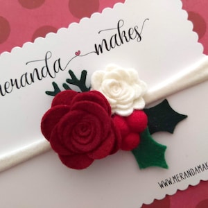 May include: A white headband with a red felt rose, a white felt rose, green felt holly leaves, and red felt berries. The headband is on a white card with the text "meranda makes" and "www.merandamakes.com".