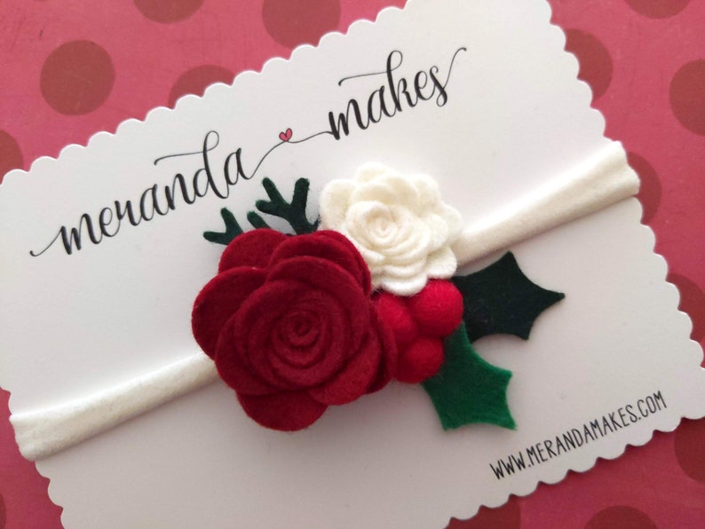 May include: A white headband with a red felt rose, a white felt rose, green felt holly leaves, and red felt berries. The headband is on a white card with the text "meranda makes" and "www.merandamakes.com".