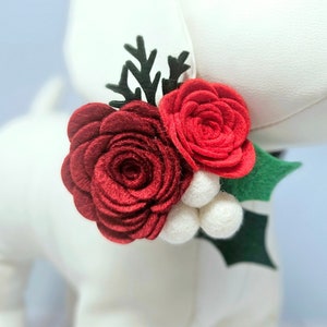 May include: A felt flower hair clip with two red roses and white felt balls. The roses are layered and have a textured appearance. The clip is decorated with a green felt leaf.
