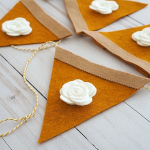 May include: A felt banner with four orange triangle flags, each with a white felt flower. The flags are connected with a white and yellow twine.