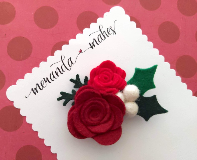 May include: Red felt roses with white felt berries and green felt holly leaves. The flowers are arranged in a cluster and are attached to a white card with the text "meranda makes" written in black cursive.